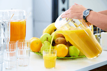 Person pours orange juice from a jug into a glass, surrounded by fresh fruit.