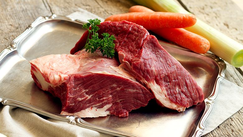 Two pieces of raw meat on a silver tray on a wooden table. Next to it, carrots and leeks.