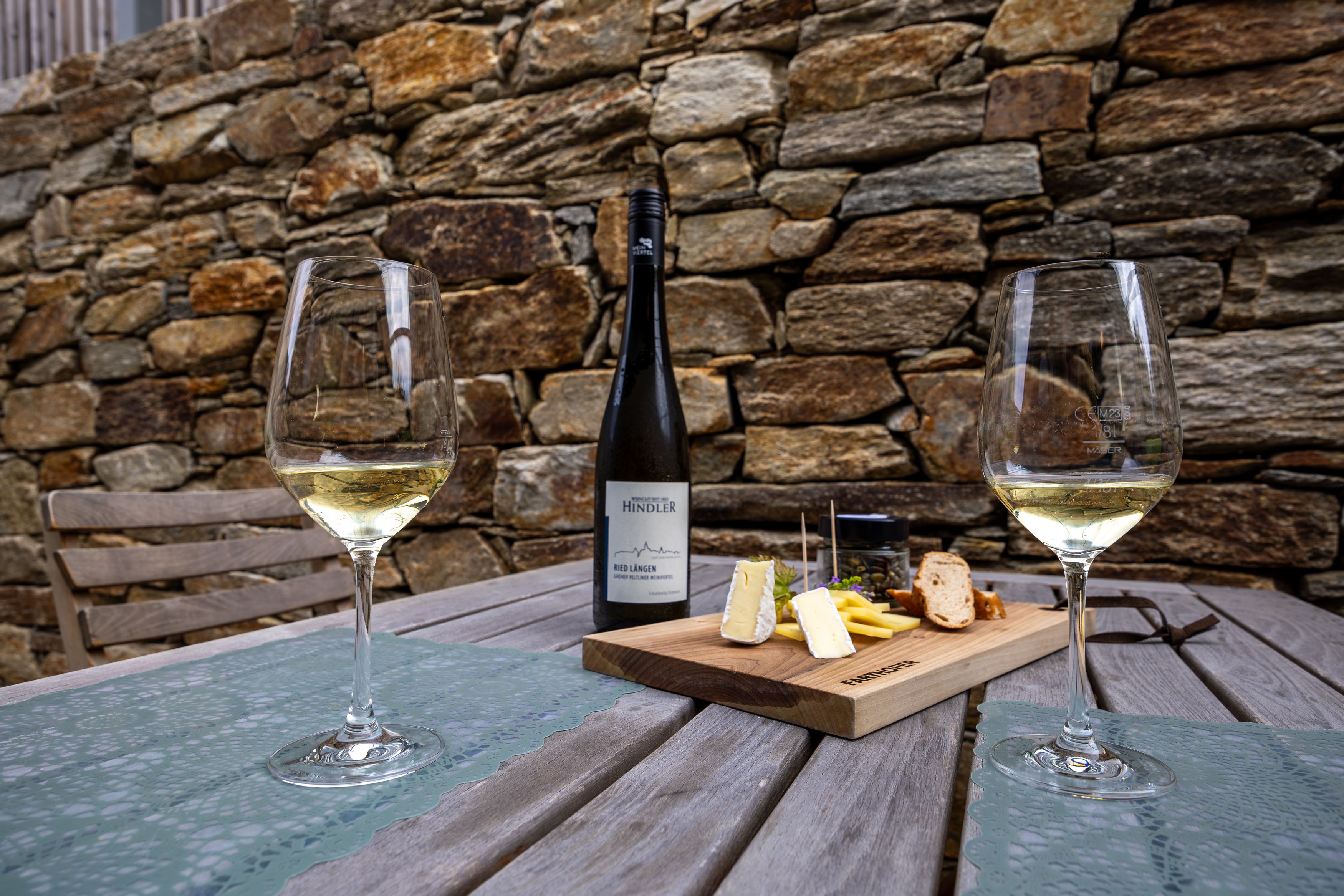 Two wine glasses and a bottle of wine on a wooden table with a cheese board, in front of a stone wall.
