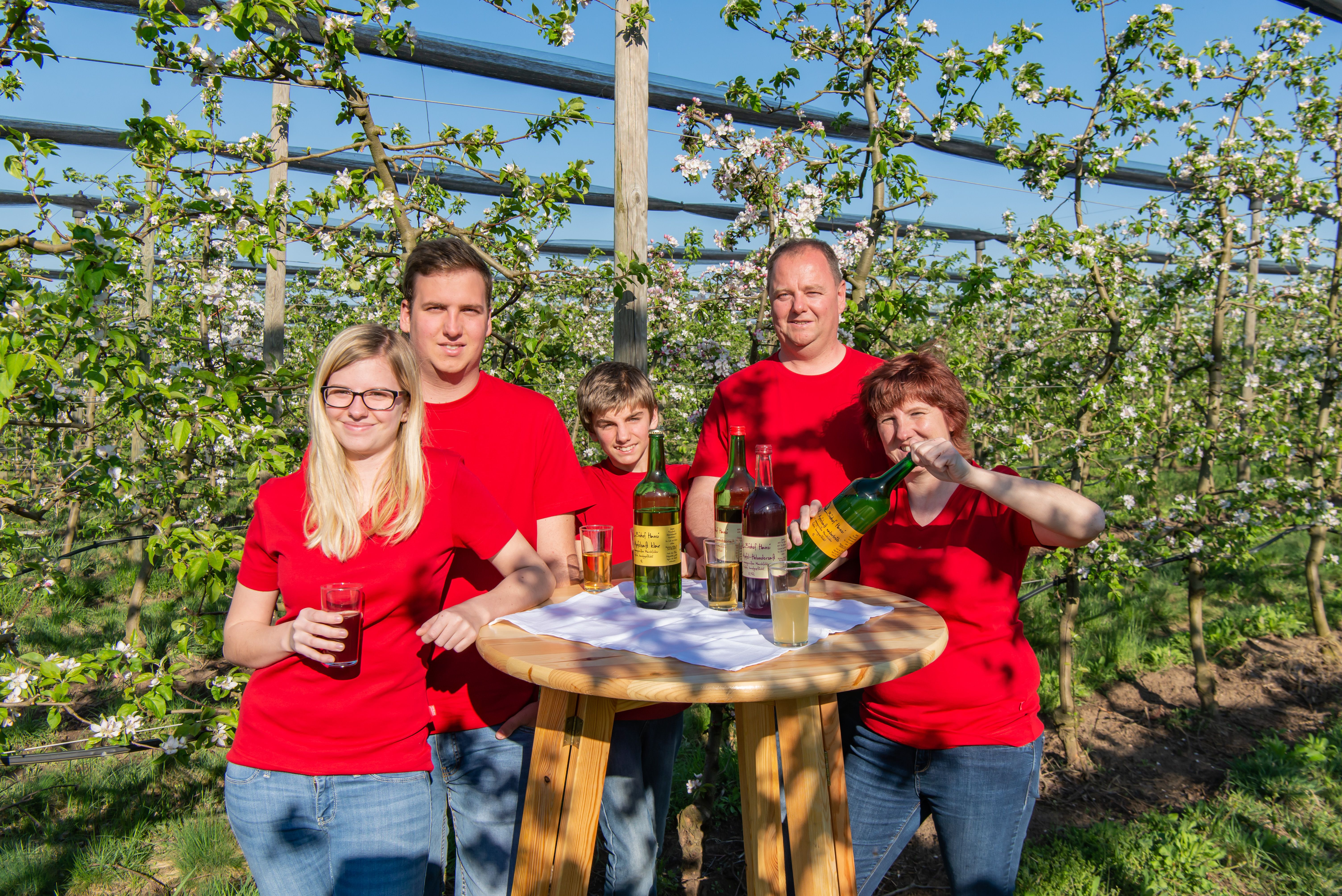A family in red T-shirts stands at a wooden table with drinks in a blossoming orchard.