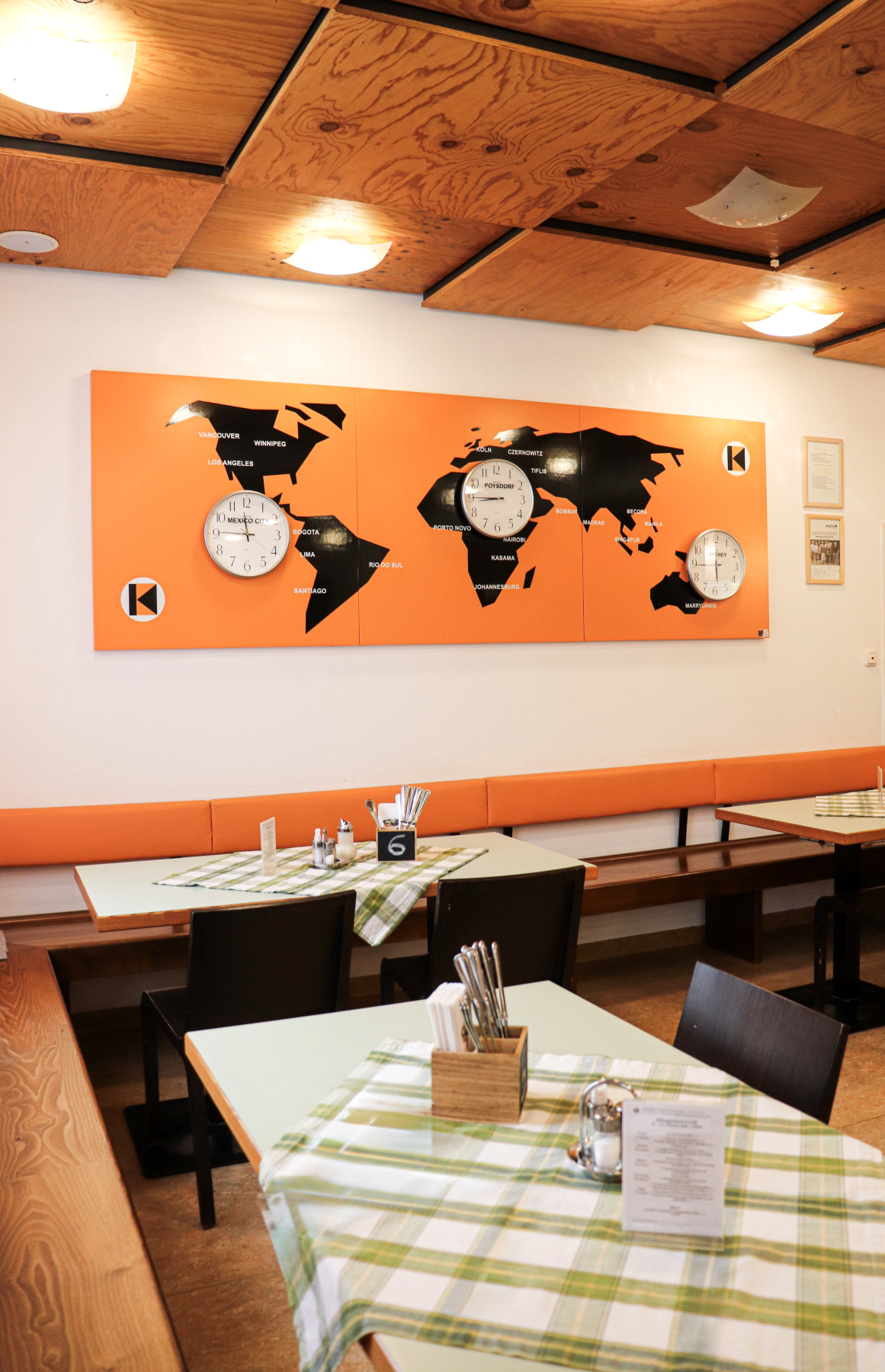 The interior of the Kolping House, featuring a ceiling with exposed wooden beams, an orange-colored wall with a map of the world, and tables with checkered tablecloths.