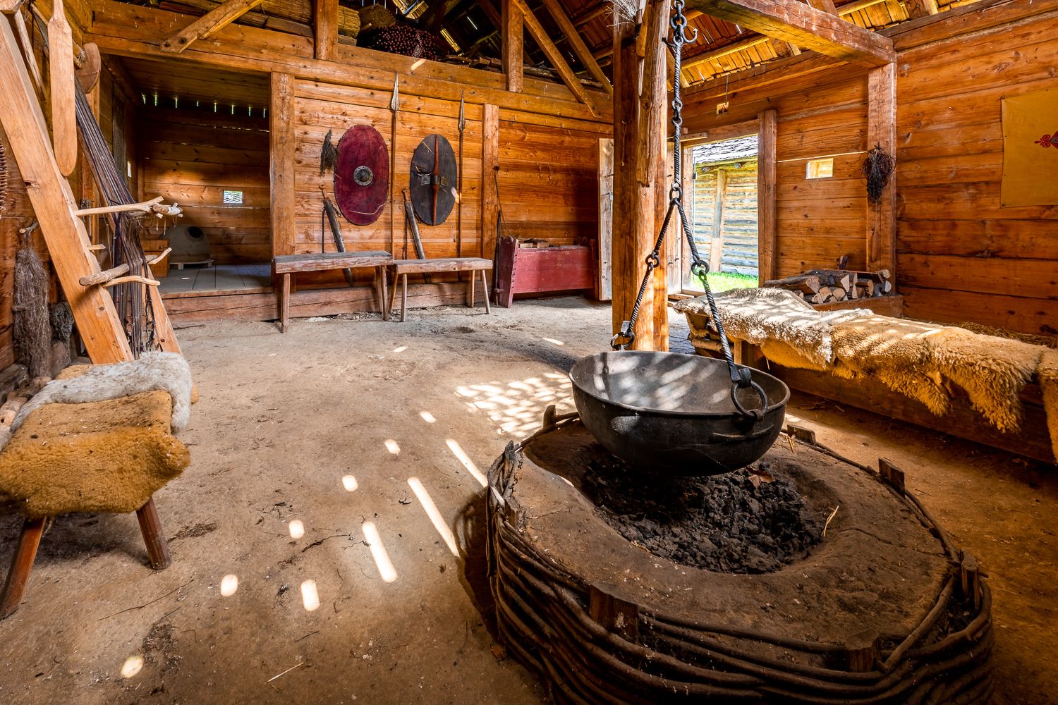 Interior view of a reconstructed Celtic house with wooden walls, a large cauldron over a fireplace and shields on the wall.
