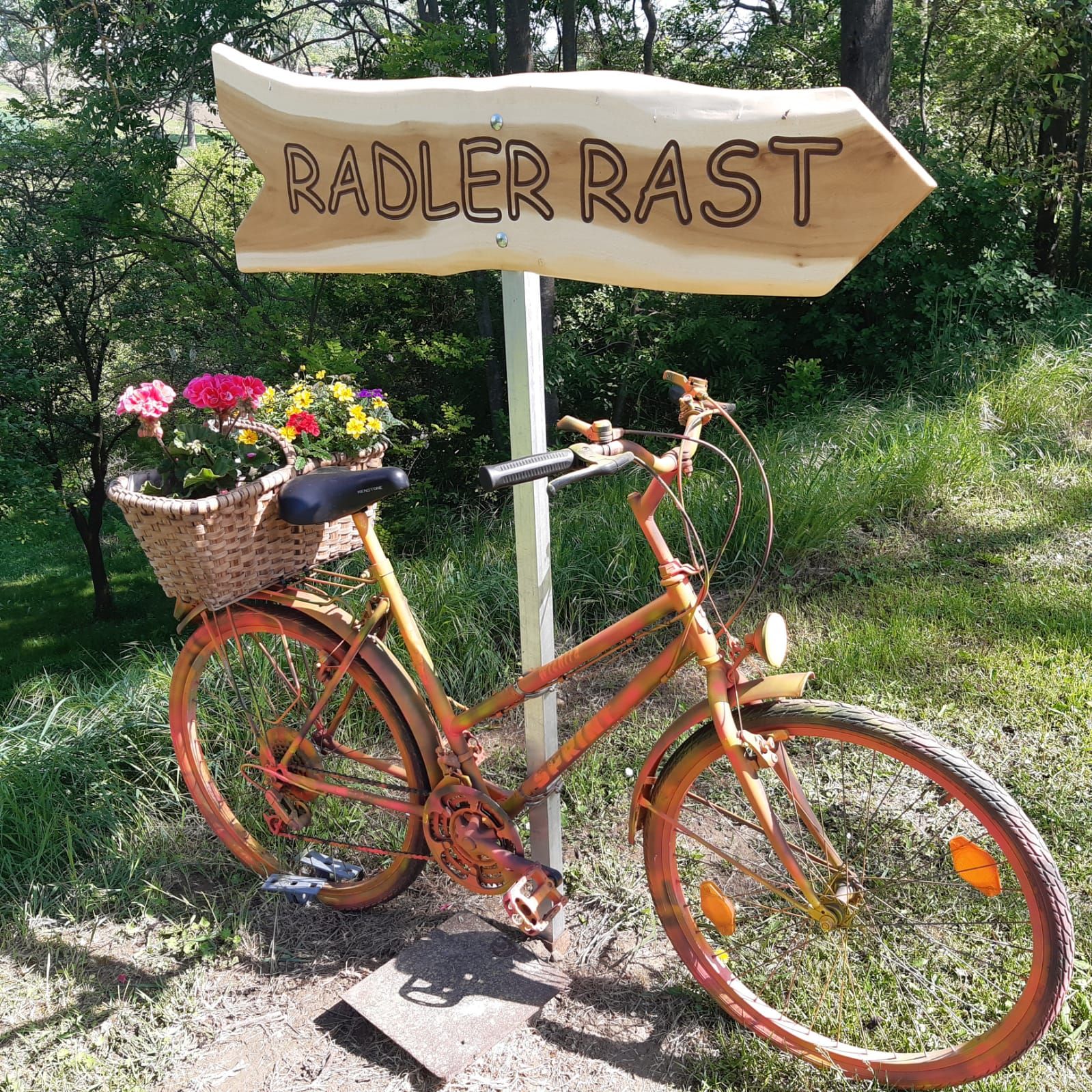 An orange bicycle with a basket of flowers stands next to a wooden sign that reads "Radler Rast" in a green setting.