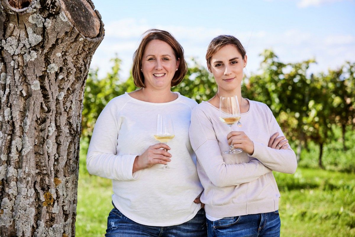 Two women stand with wine glasses in front of a tree in a vineyard.