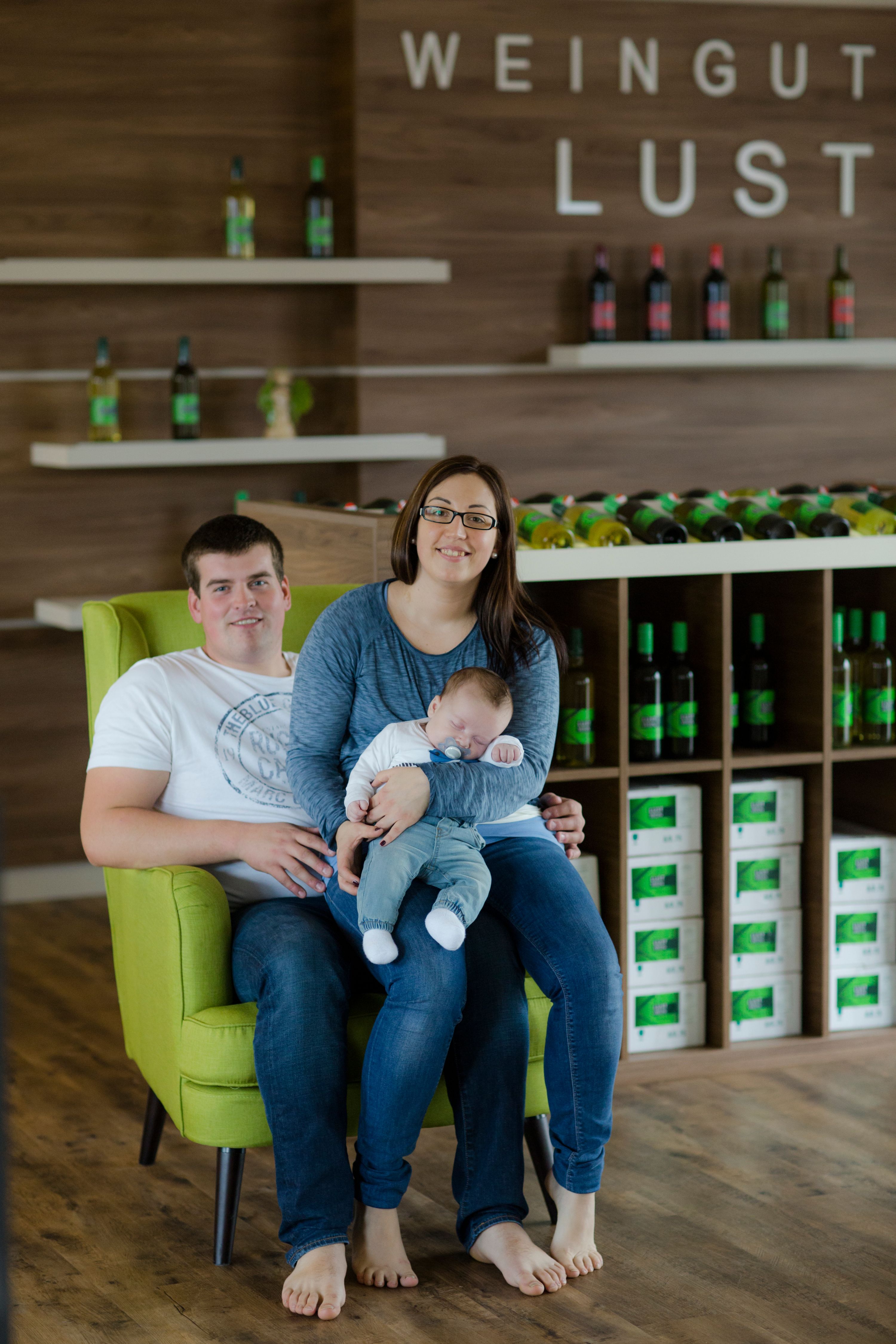 A couple with a baby sits on a green armchair in a winery.