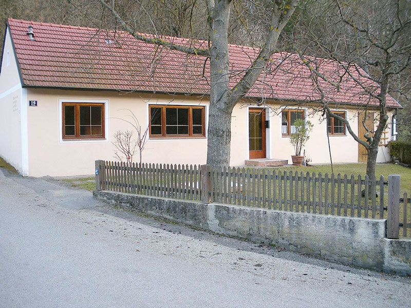 A single-storey vacation home with a red tiled roof and light-colored paint, surrounded by a wooden fence and trees.