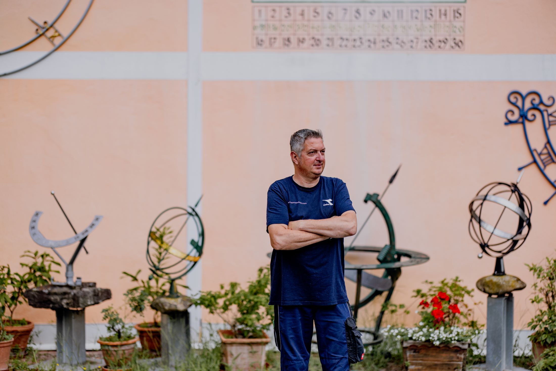A man stands with his arms folded in front of a wall with sundials and plants.