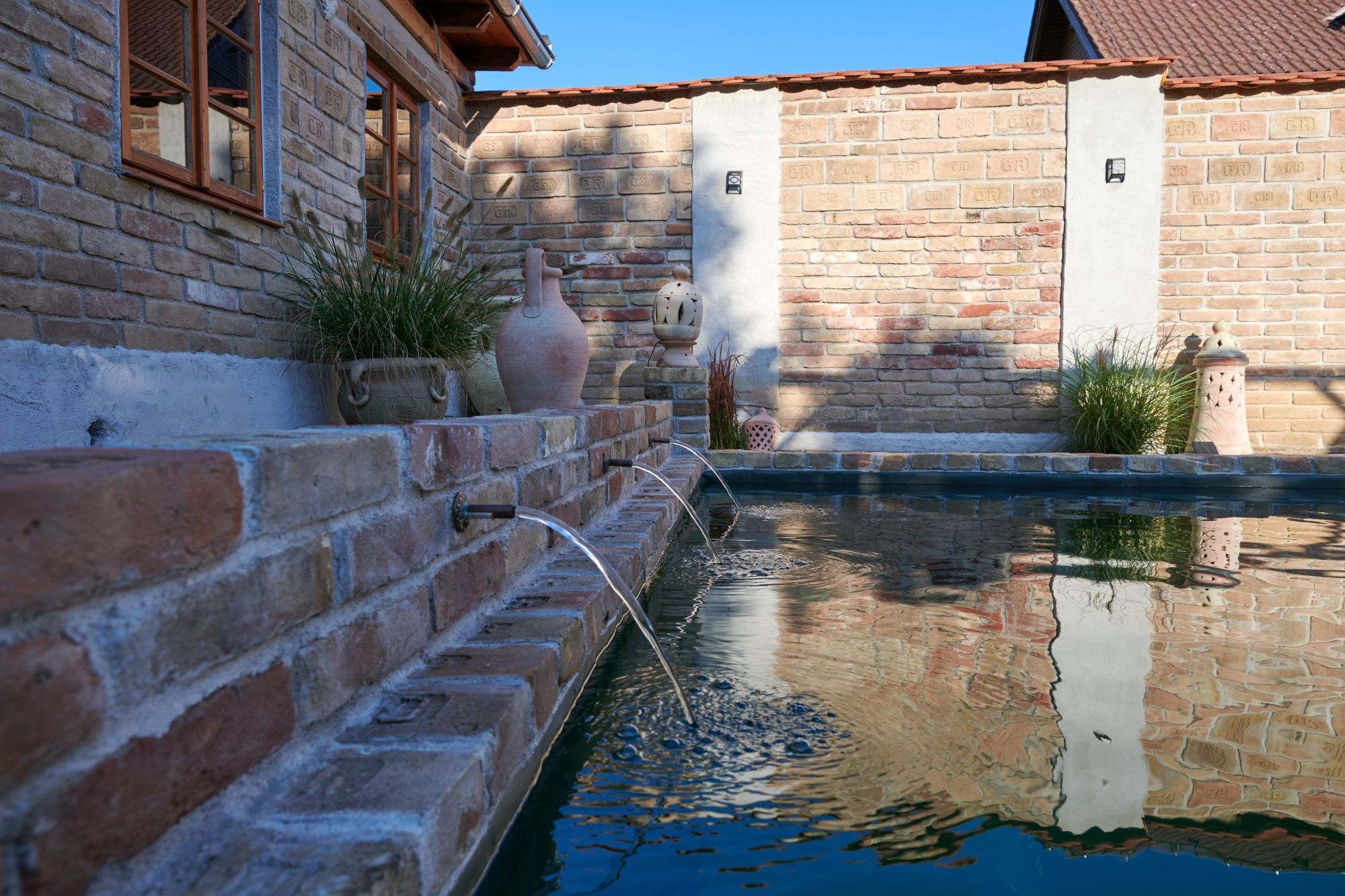 Brick wall with gargoyles and plants at the edge of a pool.