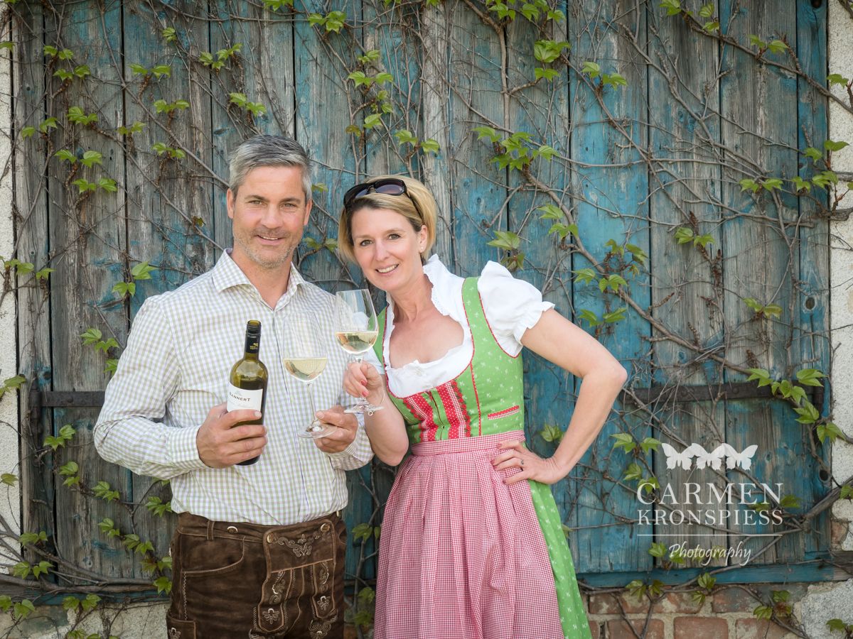 A man and a woman in traditional dress hold wine bottles and glasses in front of an overgrown wooden wall.