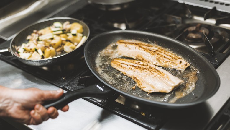 Two fish fillets fry in a pan on a stove, with a pan of vegetables next to them.