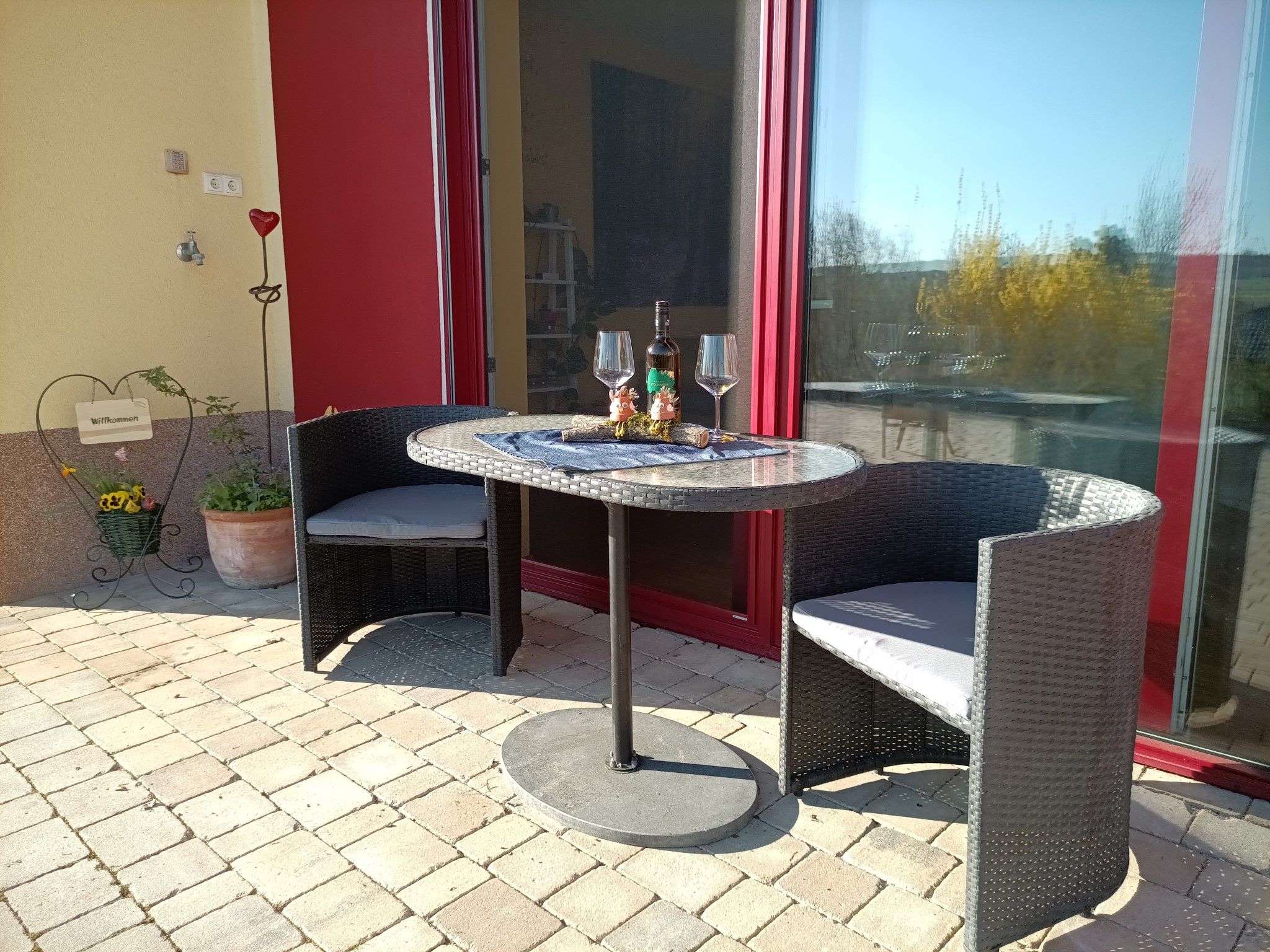 Terrace with table, two chairs, bottle of wine and glasses in front of a red door.