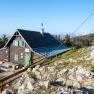 A mountain hut with green shutters and a summit cross in the foreground, surrounded by rocks and trees.