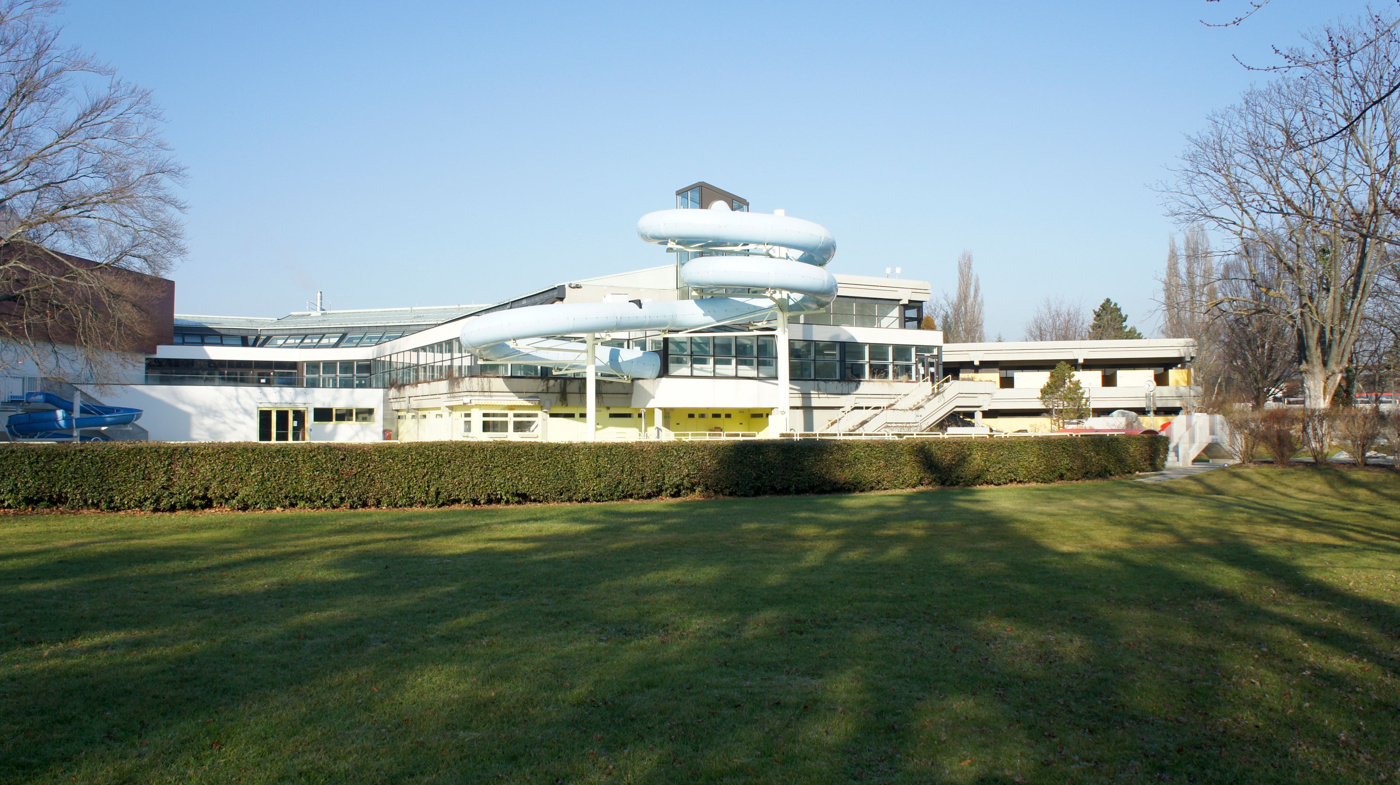 Building with blue tube slide and glass façade, surrounded by lawns and trees.