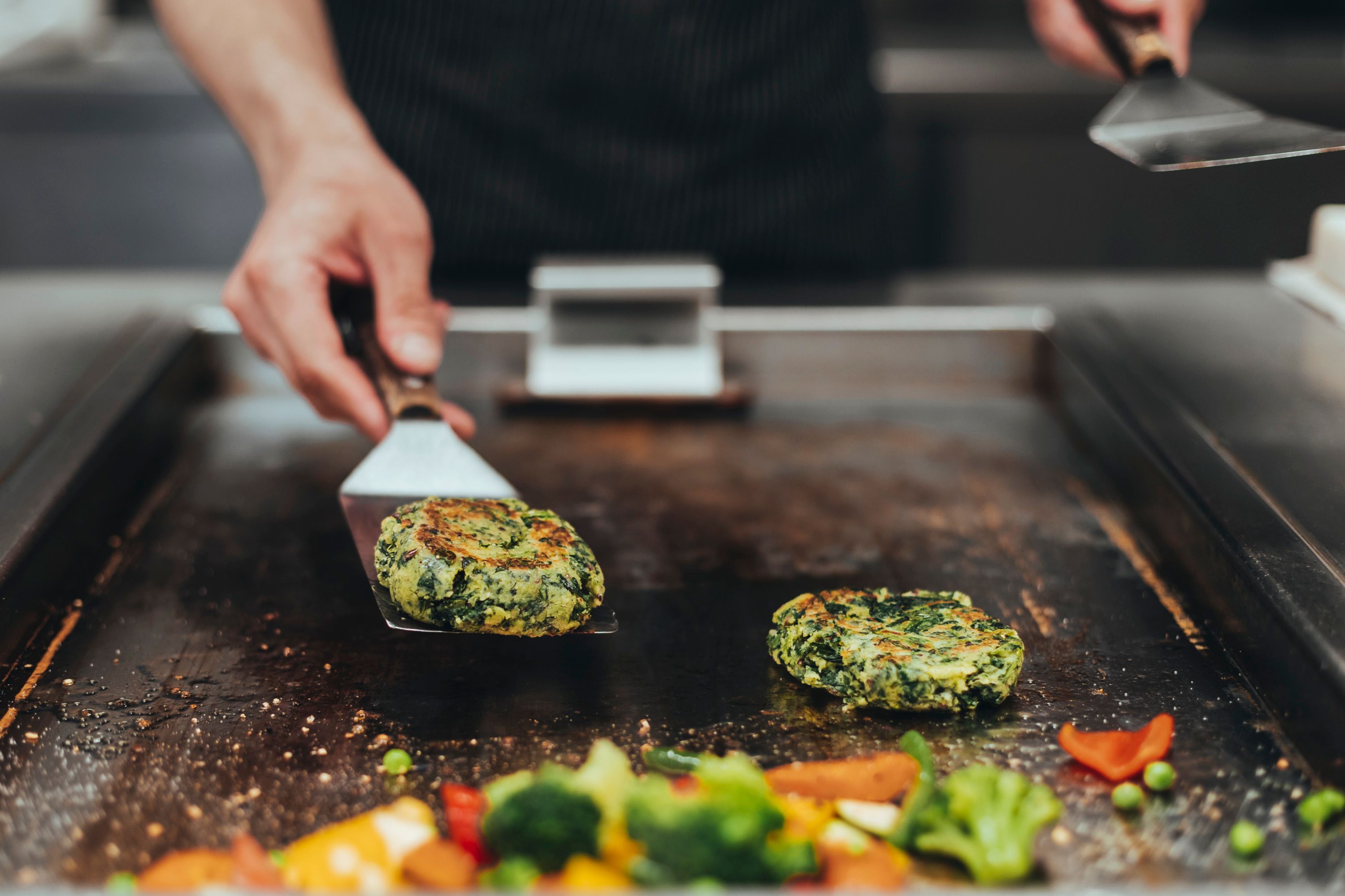 A cook turns vegetable patties on a grill plate, surrounded by colorful vegetables.