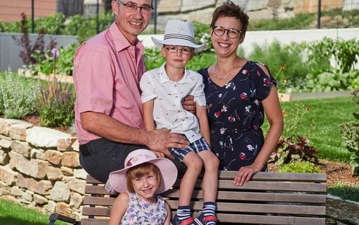 A family is sitting on a bench in the garden, consisting of two adults and two children with hats.