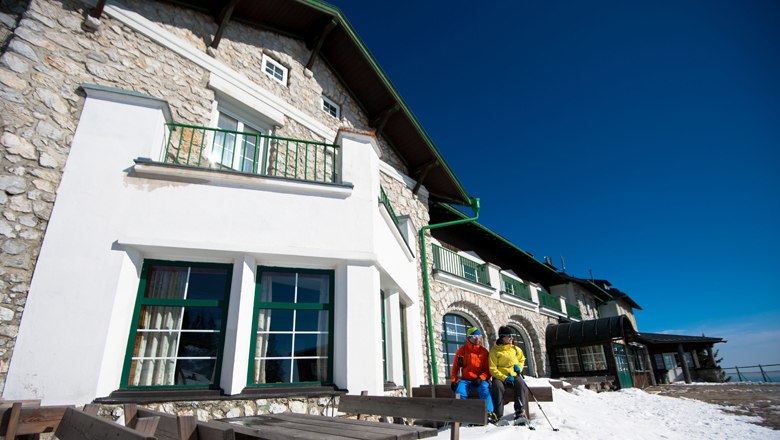 Two people in winter clothing in front of a mountain inn with snow-covered ground and blue sky.