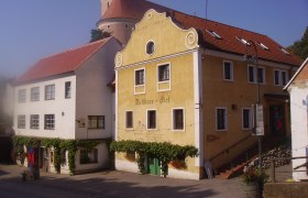 Historic building with yellow fa&ccedil;ade and red roof tiles, next to it a white building. In the background a tower with a dome.