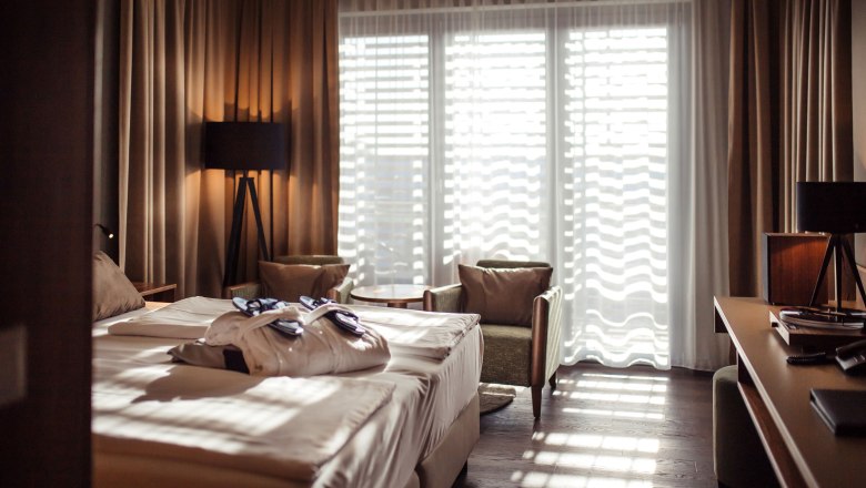 Hotel room with double bed, bathrobes, two armchairs and a table in front of a large window with curtains.