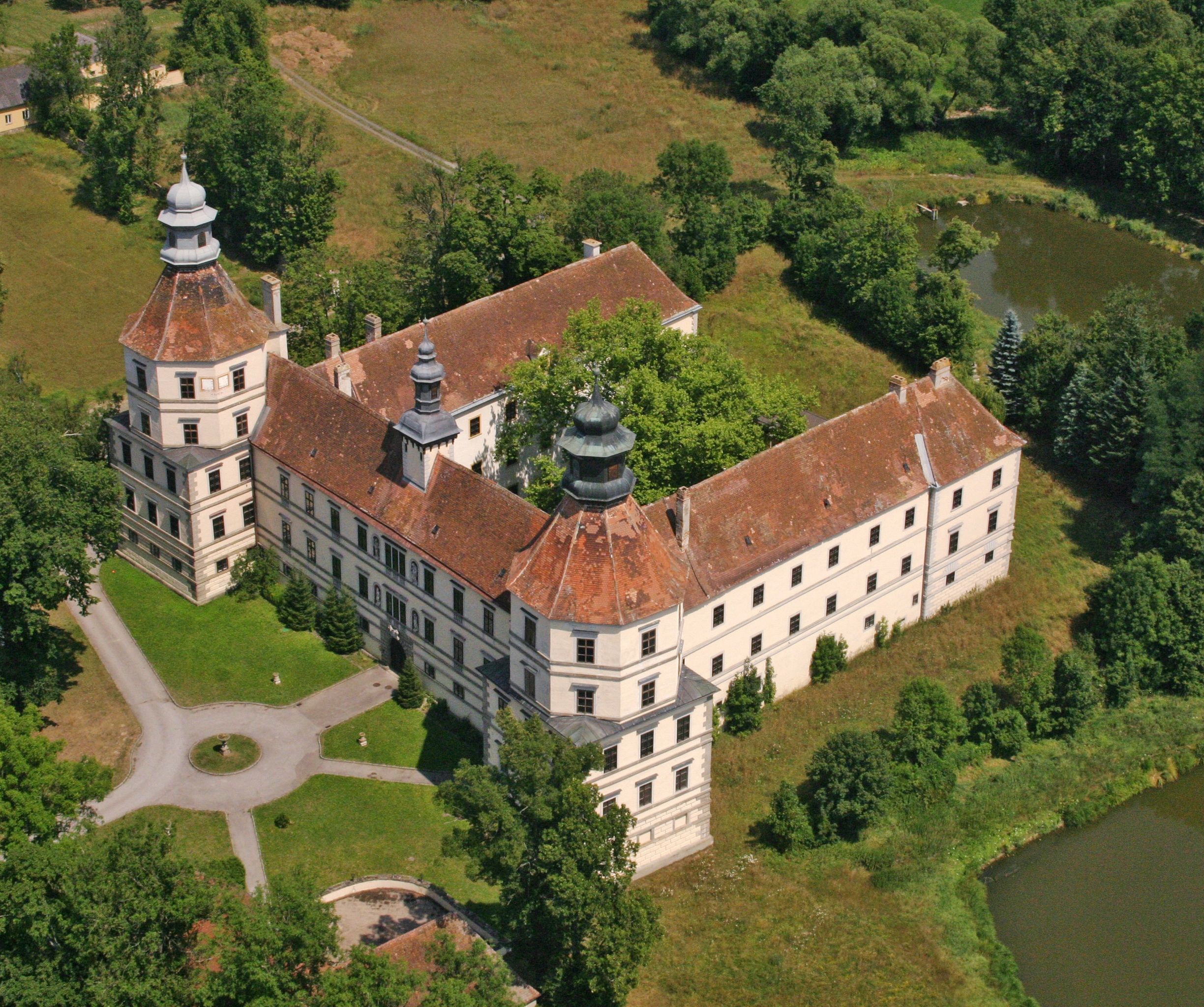 Aerial view of a historic castle with two towers and an inner courtyard, surrounded by trees and a pond.