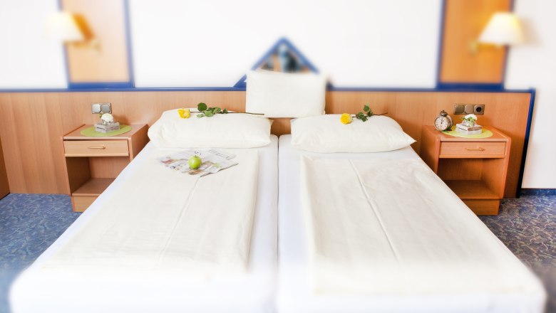 Two single beds in a hotel room with white sheets, yellow roses on the pillows and a green apple on a newspaper.