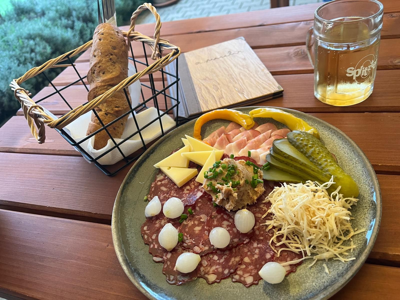 A plate with cold cuts, cheese, gherkins and onions, next to it a basket of bread and a drink on a wooden table.