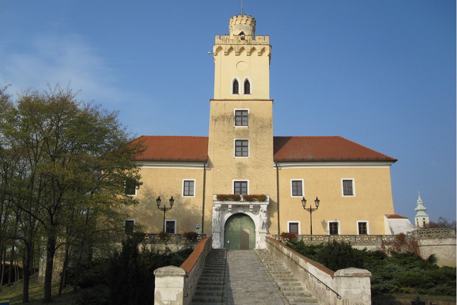 Castle with yellow façade and red roof, central tower, wide stone staircase, blue sky.