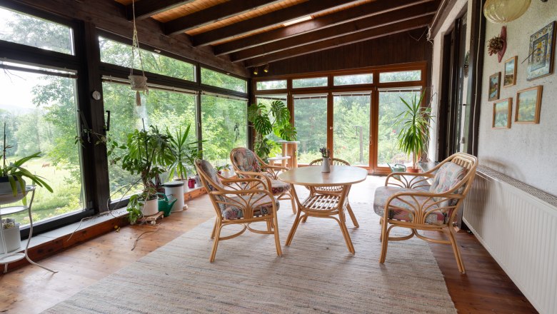 A conservatory with wooden floor, rattan furniture and lots of plants. Large windows offer a view of green nature.