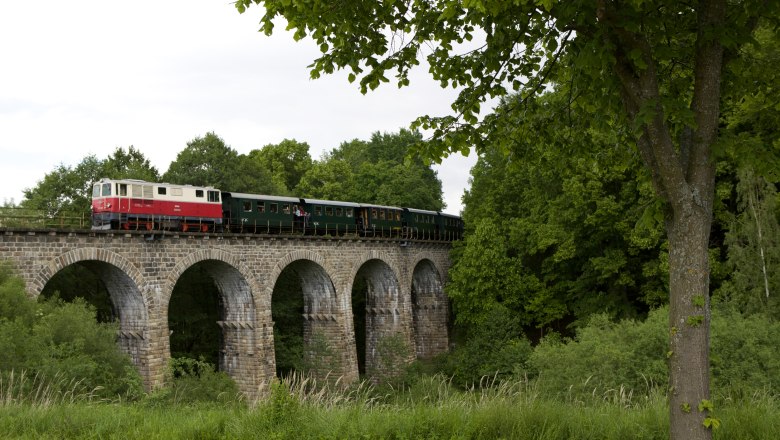 A train crosses a stone viaduct in a green landscape.
