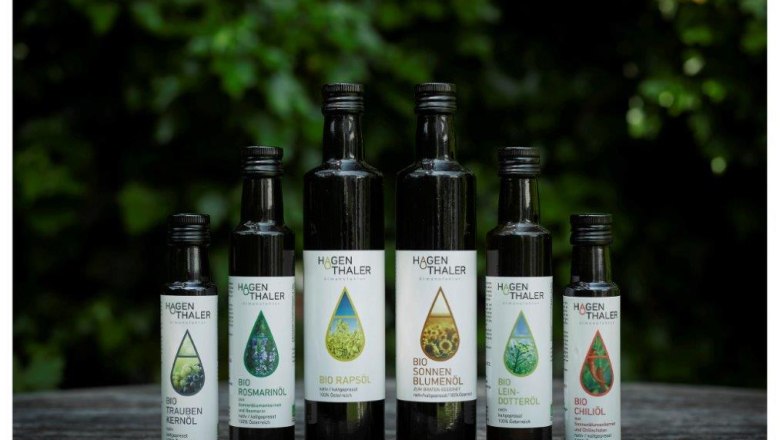 Six bottles of Hagenthaler organic oils on a wooden table against a green background.