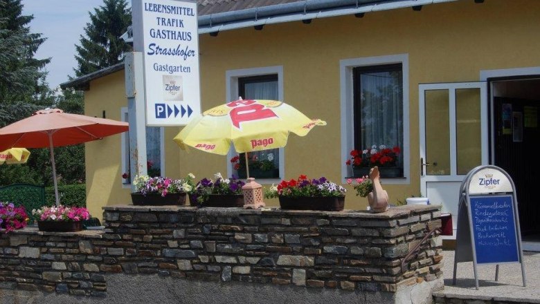 Exterior view of a yellow building with signs for food, tobacconist and inn. There are parasols and flowers in front of the house.