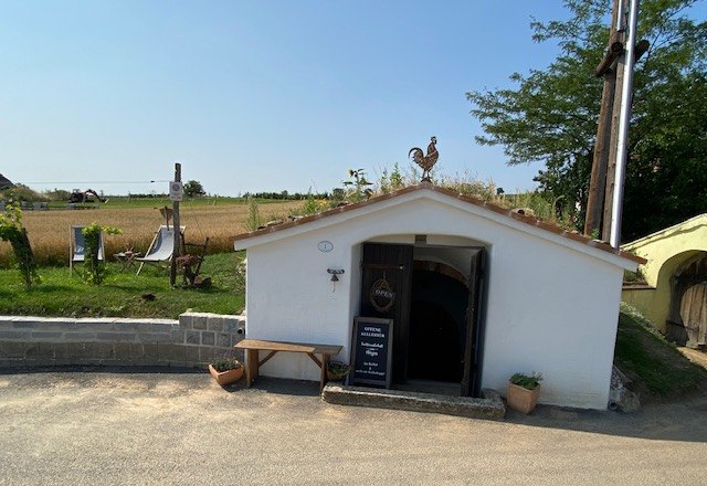 White cellar building with a rooster sculpture on the roof and an open entrance, surrounded by rural landscape.