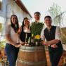 Four people in traditional dress stand around a wine barrel with flowers and bottles.