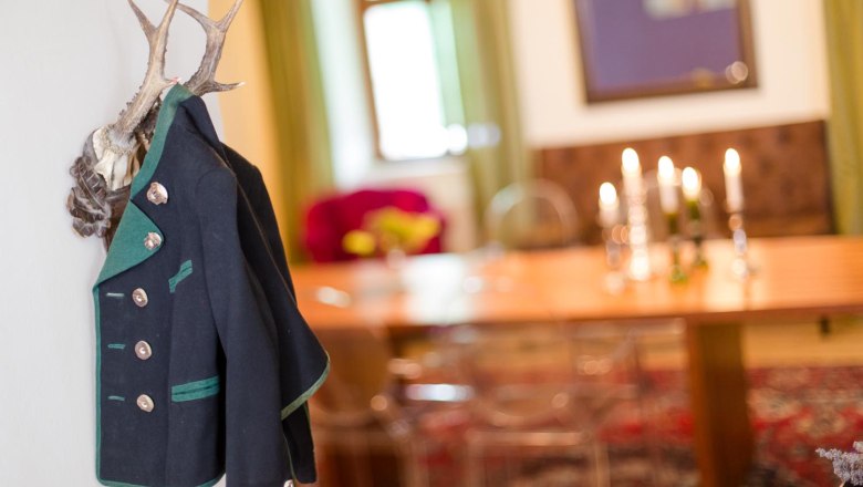 A traditional jacket hangs from antlers on the wall. In the background is a table with candlesticks and a window.
