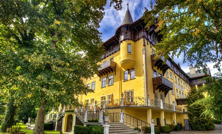Historic yellow building with towers and decorations, surrounded by trees.