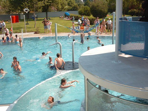 People swim and play in an outdoor pool with a slide and green area.