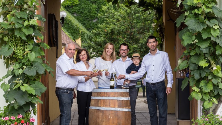 Group of people standing around a wine barrel, holding wine glasses and smiling at the camera. Surrounded by vines and flowers.