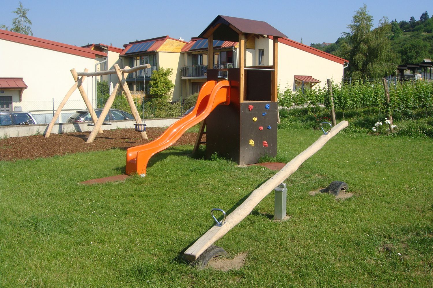 A children's playground with a slide, swing and seesaw on a meadow in front of residential buildings.