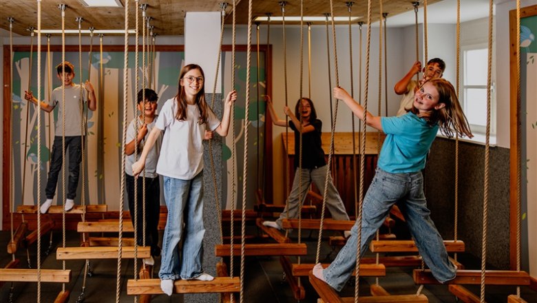 Children play on an indoor ropes course with wooden platforms.
