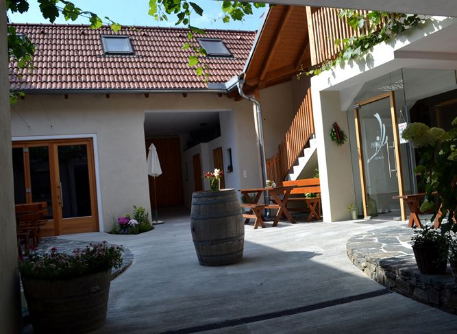 A cozy courtyard with wooden furniture, plants and a wine barrel. A building with red roof tiles and a wooden staircase in the background.