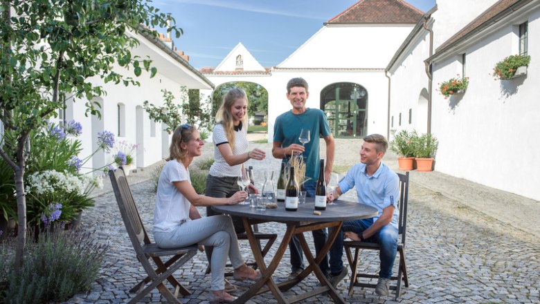 Four people sit at a round table outside and enjoy wine. They are surrounded by white buildings.