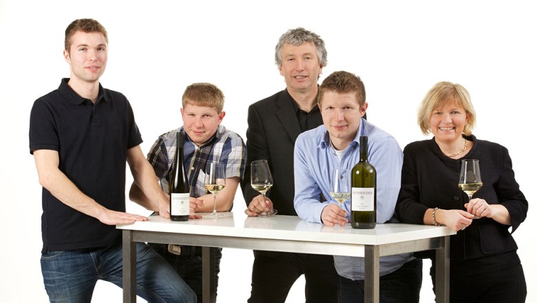 Five people are standing around a table with bottles of wine and glasses.