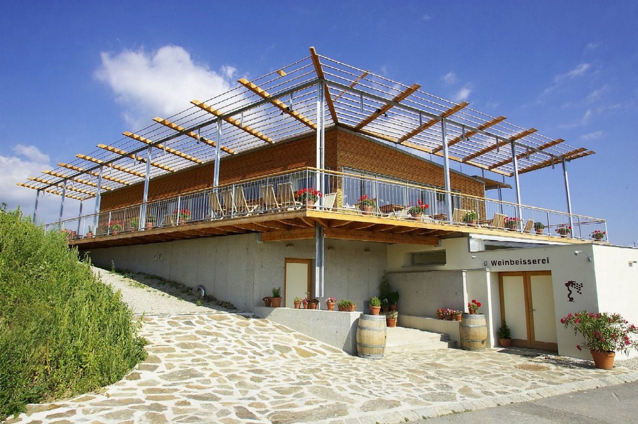 Modern building with terrace and pergola, surrounded by plants and wine barrels, under a blue sky.