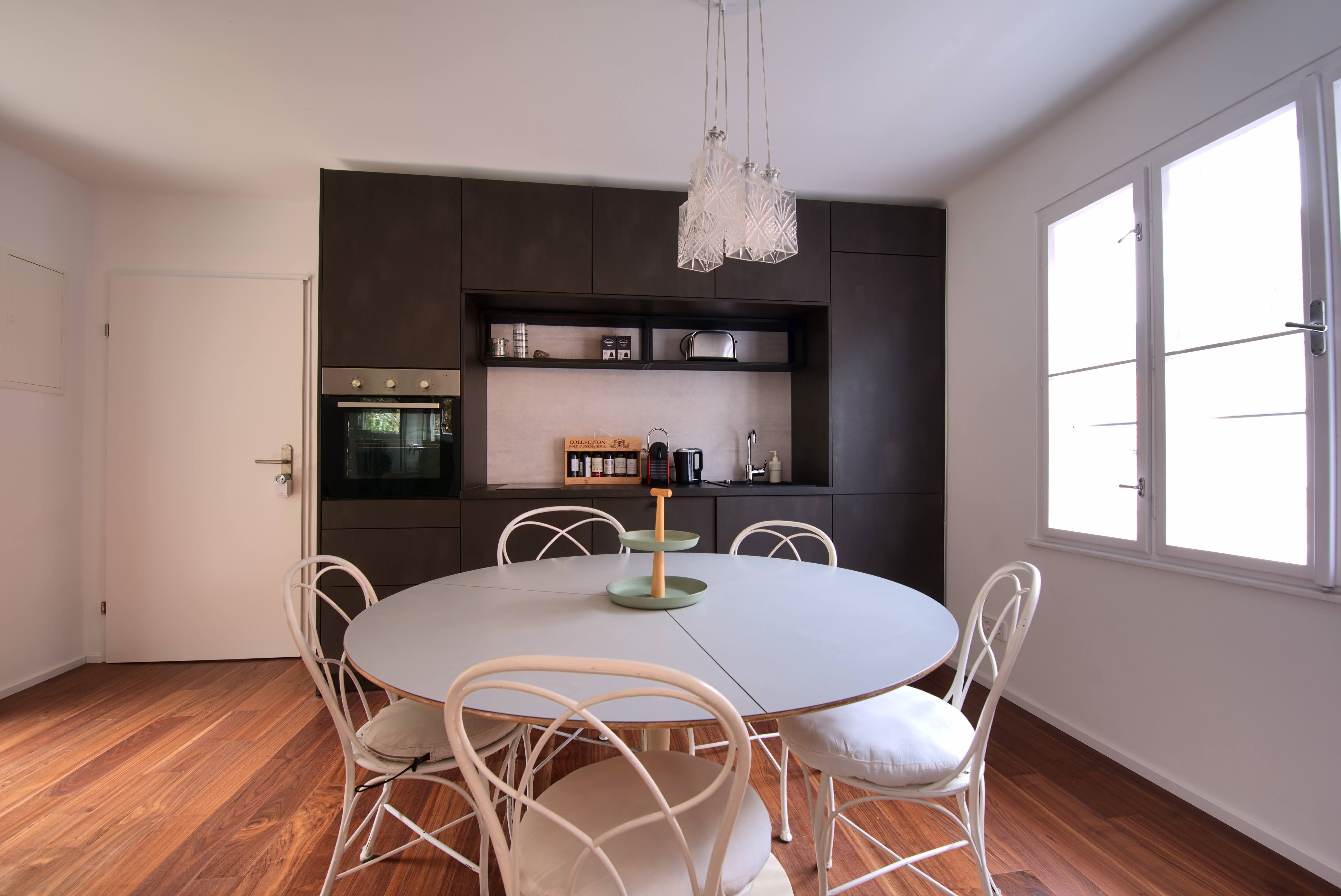Modern dining area with round table, white chairs and dark kitchen unit in the background.