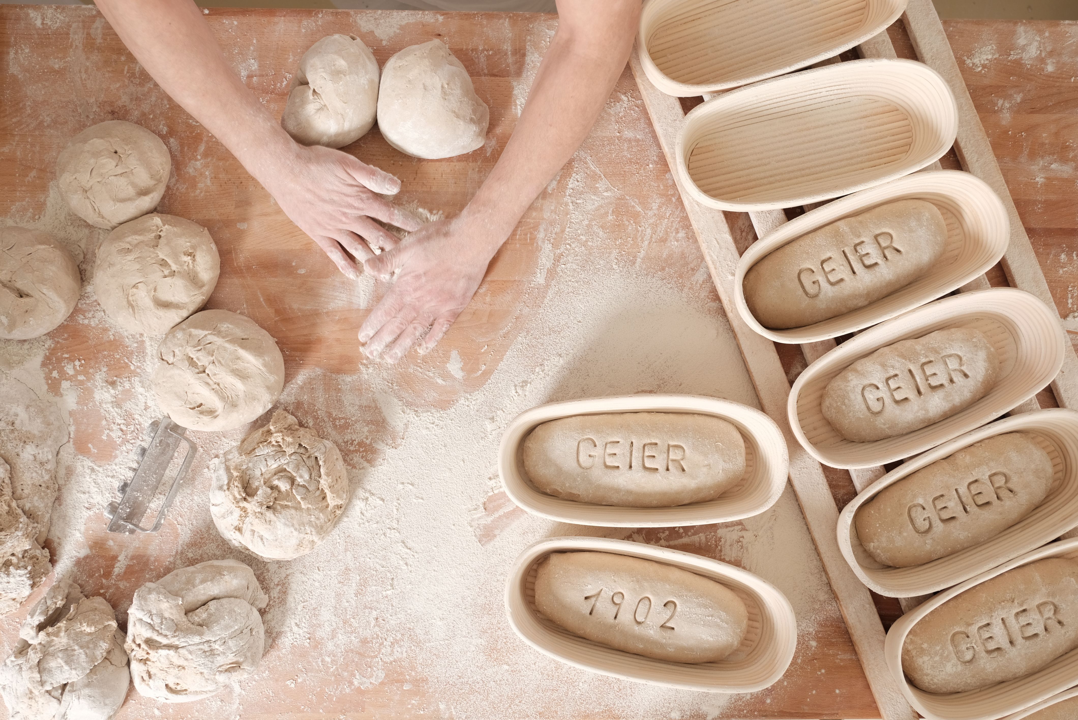 Hands form dough on a floured table, next to proofing baskets with "GEIER" and "1902".
