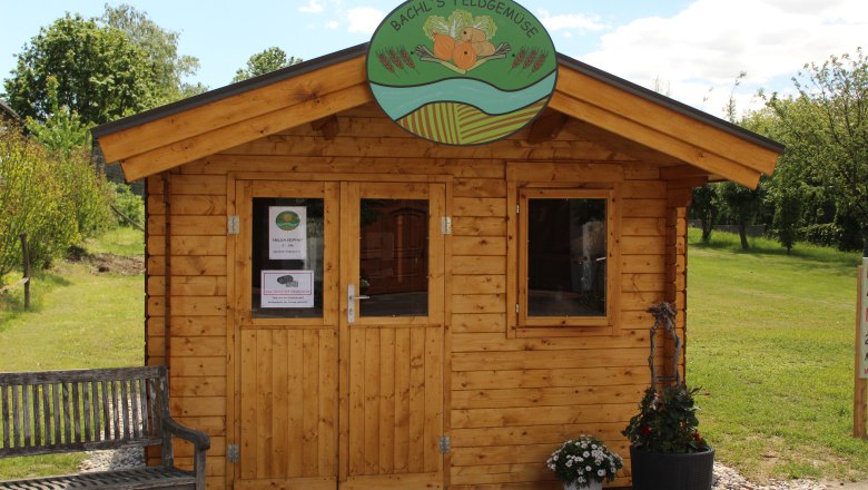 Wooden hut with sign 'Bachl's Feldgem&uuml;se' and flowers in front of it.