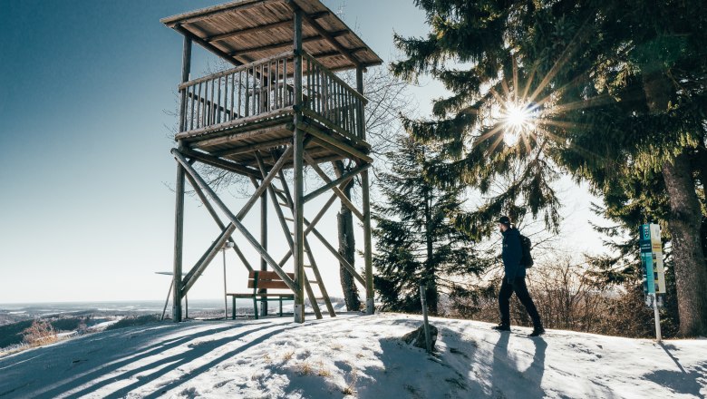 A wooden lookout post on a snowy hill with a hiker in the foreground and rays of sunshine through the trees.