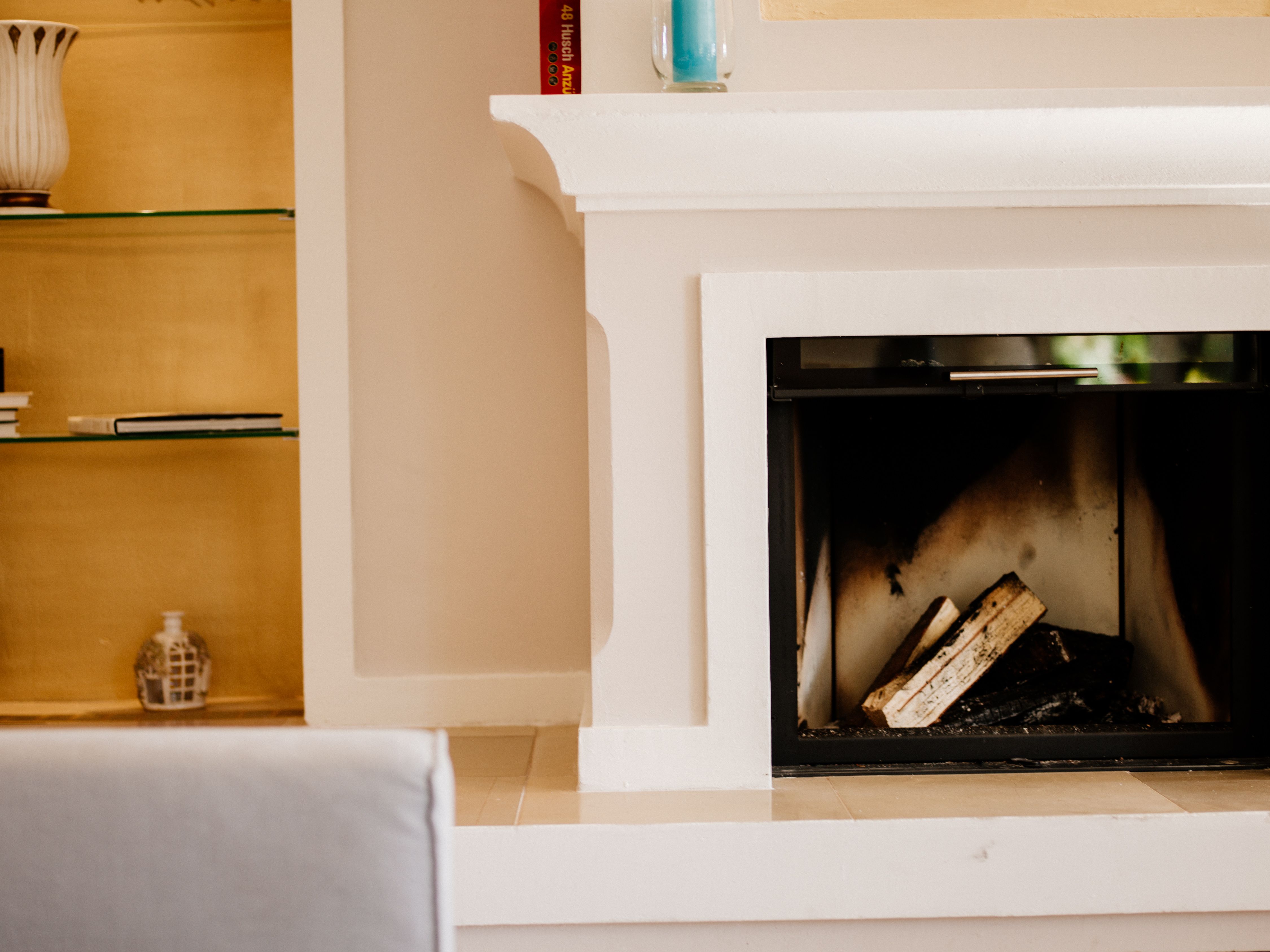 Interior view of a fireplace with logs, next to it a shelf with books and decoration.