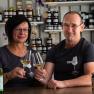 A man and a woman sit at a table in a farm store, holding glasses of wine and smiling. Shelves with products can be seen in the background.