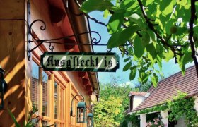 A cozy outdoor area of a wine tavern with wooden tables and flowering plants, shaded by trees.