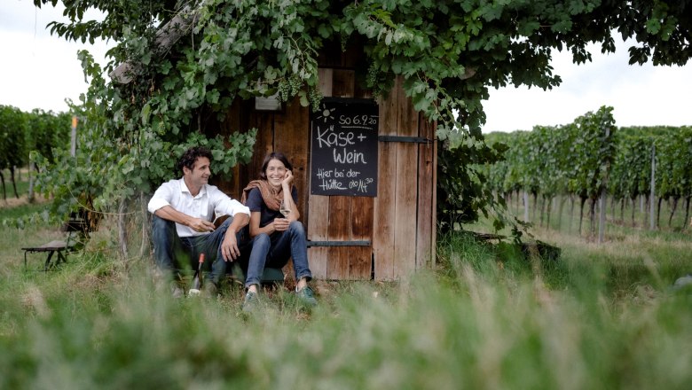 Two people are sitting in front of a wine hut in a vineyard.